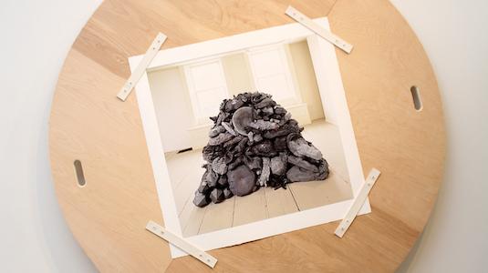 Photograph of the sculpture "Heap, Concord," a pyramidal pile of grey objects, felt taped onto a circular plywood  base.
